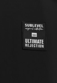Schwarzer Stoff mit einem rechteckigen Etikett, das "SUBLEVEL Originalkollektion", "GRÜNDUNG 1991" und "ULTIMATIVE ABLEHNUNG" in kontrastierendem weißen Text anzeigt.