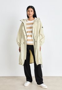 Világos bézs hosszú parkabunda kapucnival, rugalmas mandzsettákkal és zsineggel állítható derékkal, fehér és bézs csíkos póló fölött viselve. Fekete nadrág és fehér sportcipő.