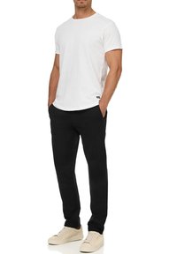 Weißes Baumwoll-T-Shirt, kurze Ärmel, runder Halsausschnitt; kombiniert mit schwarzen Hosen. Das Modell trägt beige Sneakers und hat die Hände in den Taschen. Einfaches Design.
