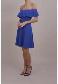 Blå av-axel klänning med en ruffled topp, åtsittande midja och utsvängd kjol. Har en slät textur och är kombinerad med svarta remsandaler.