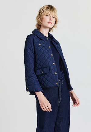 Woman with short blonde hair wearing a navy quilted jacket, dark blue top, and high-waisted dark denim jeans against a plain background.