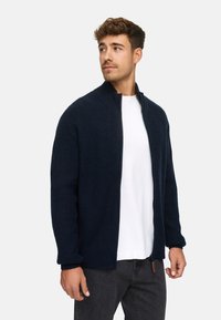 Sudadera navy con cierre y textura acanalada, cuello alto y tejido resistente. Llevada sobre una camiseta blanca, con sutiles detalles metálicos.