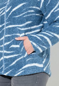 Puszysta niebieska kurtka z białymi falistymi paskami, długimi rękawami i kieszeniami bocznymi. Materiał ma miękkie, teksturowane wykończenie.