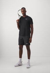 Camisa negra de Nike de manga corta con escote en V, pantalones cortos grises con el logo de Nike, sosteniendo una raqueta de tenis, usando zapatillas blancas de Nike.