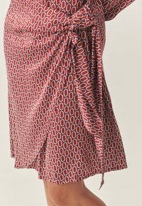 Rochie roșie cu model și un detaliu nod la talie. Prezintă un model repetitiv cu zale de lanț în alb și albastru. Material fin și fluid.