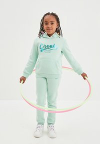 Felpa turchese chiaro con logo "Creeks", pantaloni della tuta abbinati e sneaker bianche. Bambino che tiene due hula hoop colorati.