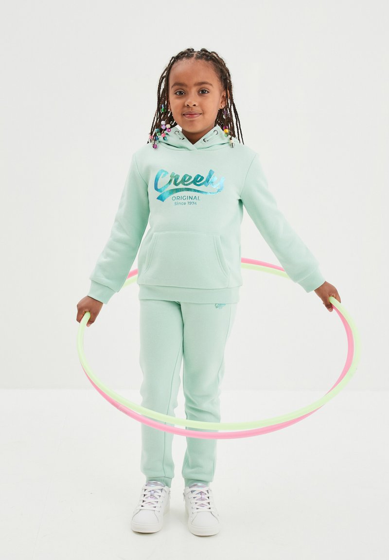 Felpa turchese chiaro con logo "Creeks", pantaloni della tuta abbinati e sneaker bianche. Bambino che tiene due hula hoop colorati.