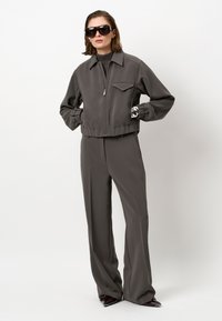 Vrouw in een donkergrijze zip-up jack en bijpassende brede pantalon, zwarte coltrui, oversized zonnebril en met een patroon bedekte handschoenen.