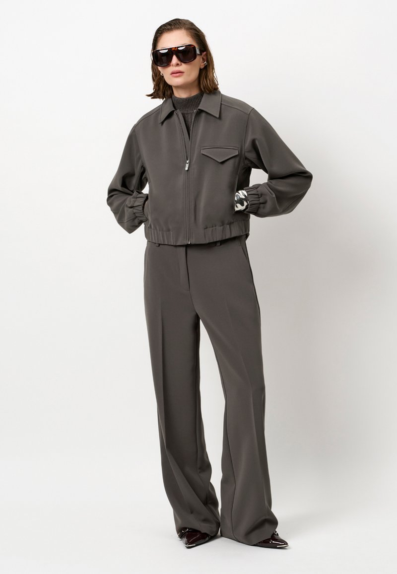 Vrouw in een donkergrijze zip-up jack en bijpassende brede pantalon, zwarte coltrui, oversized zonnebril en met een patroon bedekte handschoenen.