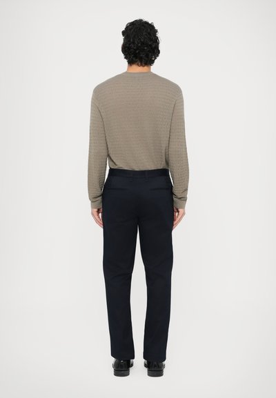 Jasnobrązowy sweter o fakturze dzianiny z okrągłym dekoltem, zestawiony z ciemnoniebieskimi spodniami, noszony z czarnymi butami, widziany od tyłu.