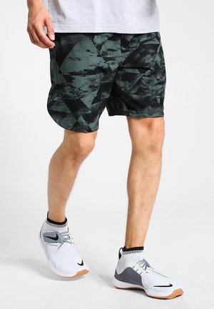 Homme portant un short à motifs géométriques noirs et gris, une chemise gris clair et des baskets blanches avec des accents noirs, en train de faire un pas en avant.