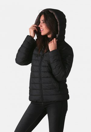 Doudoune noire avec capuche, présentant une texture matelassée et des poches zippées à l'avant, portée avec un pantalon noir décontracté.