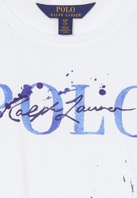 Biała koszulka z niebieskim napisem "POLO" i "Ralph Lauren" z efektem rozbryzganej farby, wykonana z bawełny. Metka widoczna wewnątrz kołnierzyka.