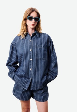 Femme portant une chemise boutonnée à rayures bleues et un short assorti, avec des lunettes de soleil bleues, les mains dans les poches, expression neutre.