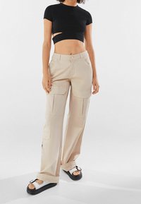 Beige cargobyxor med flera fickor, lös passform och en rosa knäppning. I kombination med en svart kortärmad ribbad topp och silverfärgade sandaler.