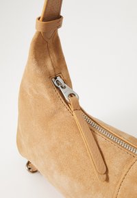 Beige semsket veske med en glatt lærreim, som har en sølvglinsende glidelås og sømdetaljer, avsmalnende form og runde metallføtter.