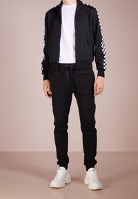 Chaqueta negra con cremallera y acentos a cuadros blancos en las mangas, combinada con pantalones jogger negros y zapatillas blancas de suela gruesa. Textura de tela suave.