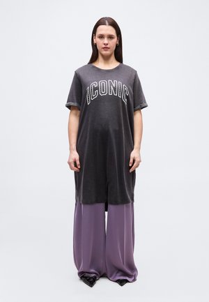 Kvinne med rett brunt hår som har på seg en løs svart kjole med teksten "ICONIC" over vide lavendelfargede bukser og svarte spisse sko, stående.