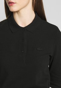 Svart polo skjorte laget av teksturert stoff, med krage, tre knapper, og en brodert Lacoste-logo i svart på venstre bryst.