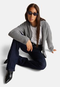 Cardigan gris de punto sobre una camiseta blanca, jeans acampanados azul oscuro y tacones negros de punta. Los complementos incluyen un collar de cadena y gafas de sol.