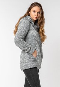 Light gray fuzzy hoodie with a textured exterior, front pockets, and a high collar, worn over dark jeans. Soft fabric enhances comfort.