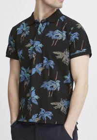 Svart polo-skjorte med et heltrykk av palmer i blå og dempede brune toner. Korte ermer og krage.