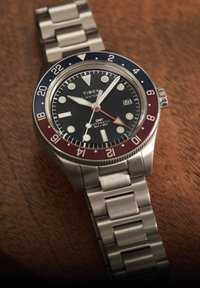 Ρολόι Timex GMT από ανοξείδωτο ατσάλι με μπλε και κόκκινη στεφάνη, μαύρο καντράν, φωσφορίζοντες δείκτες, ένδειξη ημερομηνίας και μπρασελέ από μεταλλικούς κρίκους πάνω σε ξύλινη επιφάνεια.