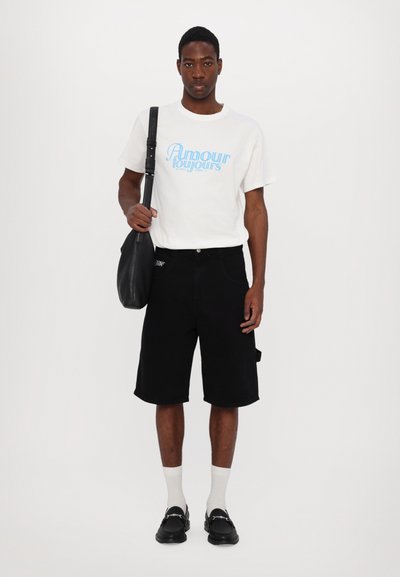 Weißes T-Shirt mit blauem Text, schwarze Jeansshorts, schwarze Tasche, weiße Socken und schwarze Slipper. Einfaches, lässiges Outfit mit minimalen Akzenten.