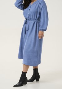 Blauwe shirtjurk met een tailleband, lange mouwen, knoopsluiting aan de voorkant en een ontspannen pasvorm; gecombineerd met zwarte enkelboots. Zachte textuur.