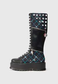 Knee-high čierne a teal kárované čižmy s kovovými cvočkami, zipsami a platformovou podrážkou. Obsahujú strieborné šnúrky a dekoratívne prívesky.