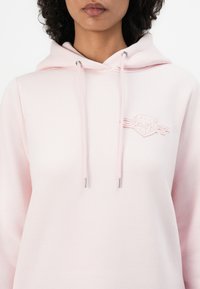 Sudadera de color rosa claro hecha de tela suave, con un bolsillo frontal, cordones ajustables con extremos de metal plateado y un logo grabado en el pecho.