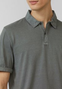 Graues Polohemd mit weicher Textur, Kragen und einer Knopfleiste mit drei Knöpfen. Aufgeschlagene Ärmel und dezente Nähte.