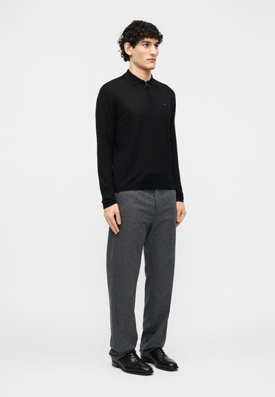 Άνδρας που στέκεται φορώντας μαύρο πόλο με μακριά μανίκια και φερμουάρ, γκρι ραμμένα παντελόνια και μαύρα δερμάτινα επίσημα παπούτσια, μπροστά σε απλό φόντο.