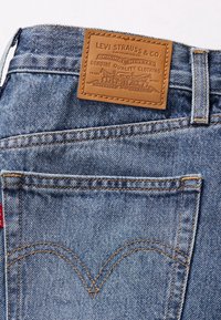 Modré džíny z rifloviny s hnědou koženou nášivkou s vyraženým textem. Viditelné švy se signaturním zakřiveným designem na zadní kapse.