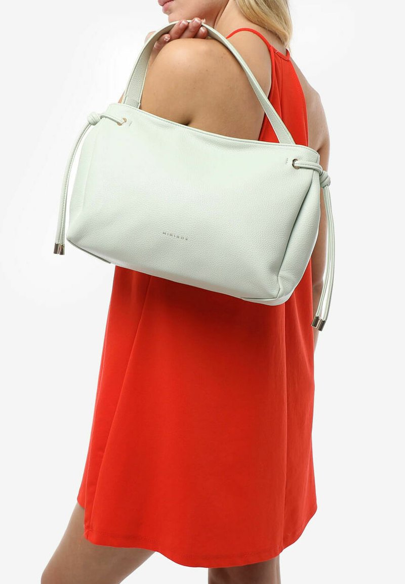 Borsa a spalla verde chiaro, con texture, dotata di chiusura a coulisse e dettagli metallici. Indossata a spalla, abbinata a un vestito arancione brillante.