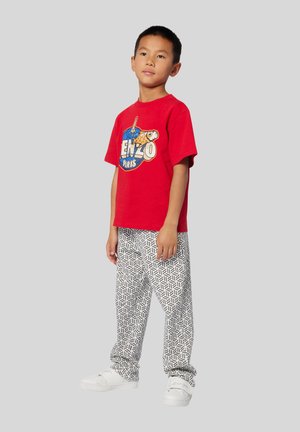 Rotes Baumwoll-T-Shirt mit Grafikdruck und Logo, kombiniert mit weißen gemusterten Hosen und weißen Sneakers. Einfaches, lässiges Outfit.