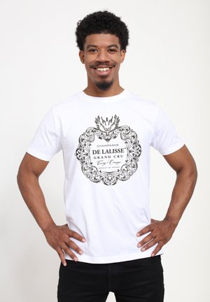 Lächelnder Mann mit Afro-Frisur trägt weißes T-Shirt mit der Aufschrift „Champagne De Lalisse Grand Cru“ und steht mit den Händen in der Hüfte vor einfarbigem Hintergrund.