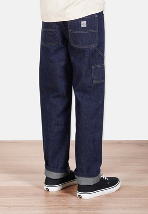 Dark blue denim cargo pants with rolled hems, back pockets, and yellow stitching. Paired with black sneakers featuring white soles.