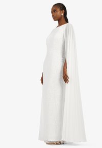 White floor-length gown with textured beading, a flowing cape detail, and a sleek silhouette. Features a boat neckline and subtle shimmer.