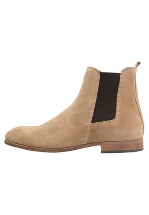 Beige ruskind Chelsea-støvle med elastiske sidepaneler, afrundet tå, træhæl og minimale syningsdetaljer. Glat tekstur over det hele.