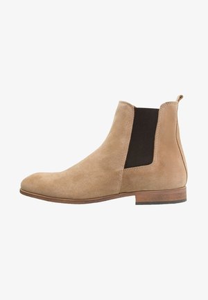 Botte Chelsea en daim beige avec panneaux latéraux élastiques, bout arrondi, talon en bois et détails de couture minimalistes. Texture lisse sur toute la surface.