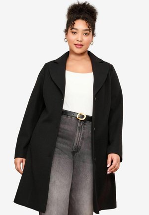 Veste noire avec un col cranté, présentant un design épuré et des boutons. Portée sur un haut blanc et associée à un pantalon gris taille haute.