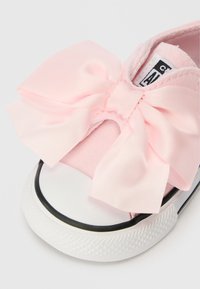 Světle růžové plátěné tenisky s velkým saténovým mašlí vpředu, bílou gumovou podešví s černým lemem a texturovaným svrchním materiálem.