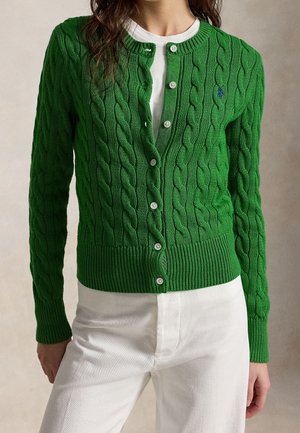 Femme portant un cardigan vert en maille torsadée boutonné sur une chemise blanche avec un pantalon blanc, debout devant un fond neutre.