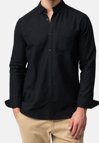 Chemise noire à manches longues avec un col à boutons, une poche poitrine et des boutons noirs. Le tissu semble lisse et légèrement texturé.