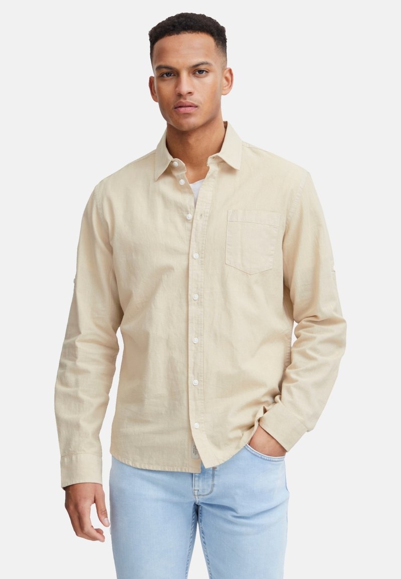 Beige Hemd mit Knopfleiste aus weichem Stoff, mit einer Brusttasche, langen Ärmeln und einem leicht abgerundeten Saum. Über einem weißen T-Shirt getragen.