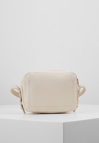 Beige kožená kabelka přes tělo s obdélníkovým tvarem, zipovým uzávěrem a dekorativními bočními smyčkami. Hladký povrch a prošití.