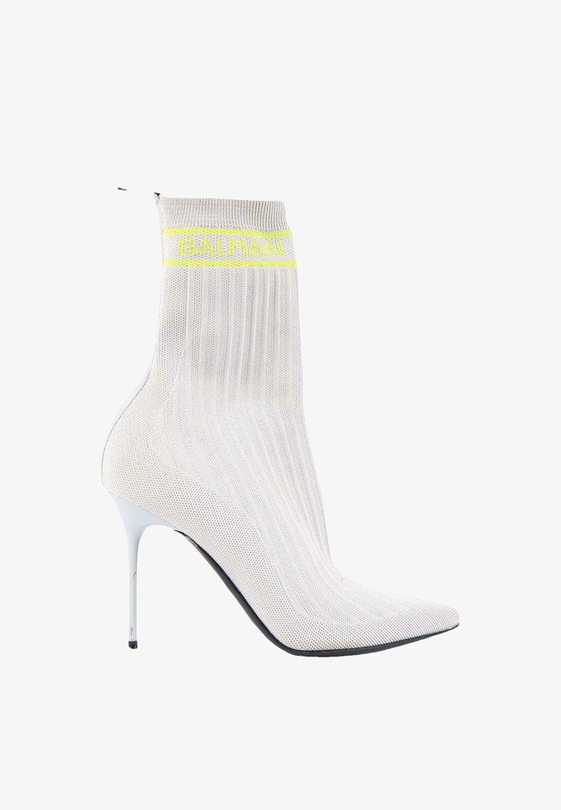 Botte à talon haut style chaussette en maille blanche avec bout pointu, talon aiguille, et logo Balmain jaune près du bord supérieur.