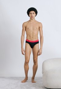 Μαύρα εσώρουχα με κόκκινη ελαστική ζώνη στη μέση. Το μοντέλο στέκεται ξυπόλητο μπροστά σε φωτεινό φόντο, με λεία υφή δέρματος και απλή στάση.