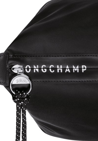 Schwarze Nylon-Tasche mit einem Reißverschluss, akzentuiert mit einem runden Metall-Zipper und dem markierten weißen Text "ONGCHAMP". Strukturiert mit einer subtilen Glanzoberfläche.